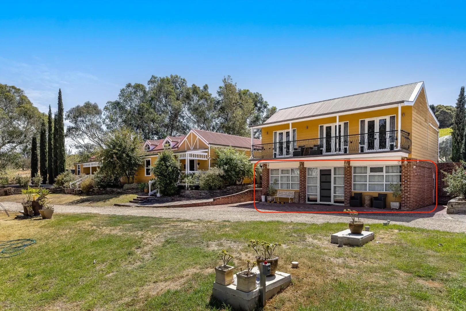 A two-story yellow house with a balcony, surrounded by a grassy yard and trees.