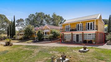 A two-story yellow house with a balcony, surrounded by a grassy yard and trees.
