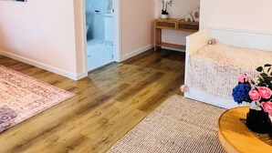 Quarto com piso de madeira, uma cama, mesa de cabeceira com um vaso de flores, espelho na parede e uma porta que dá para outro quarto.