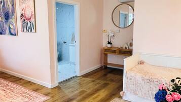A room with a wooden floor, a bed, a bedside table with a vase of flowers, a mirror on the wall, and a door leading to another room.