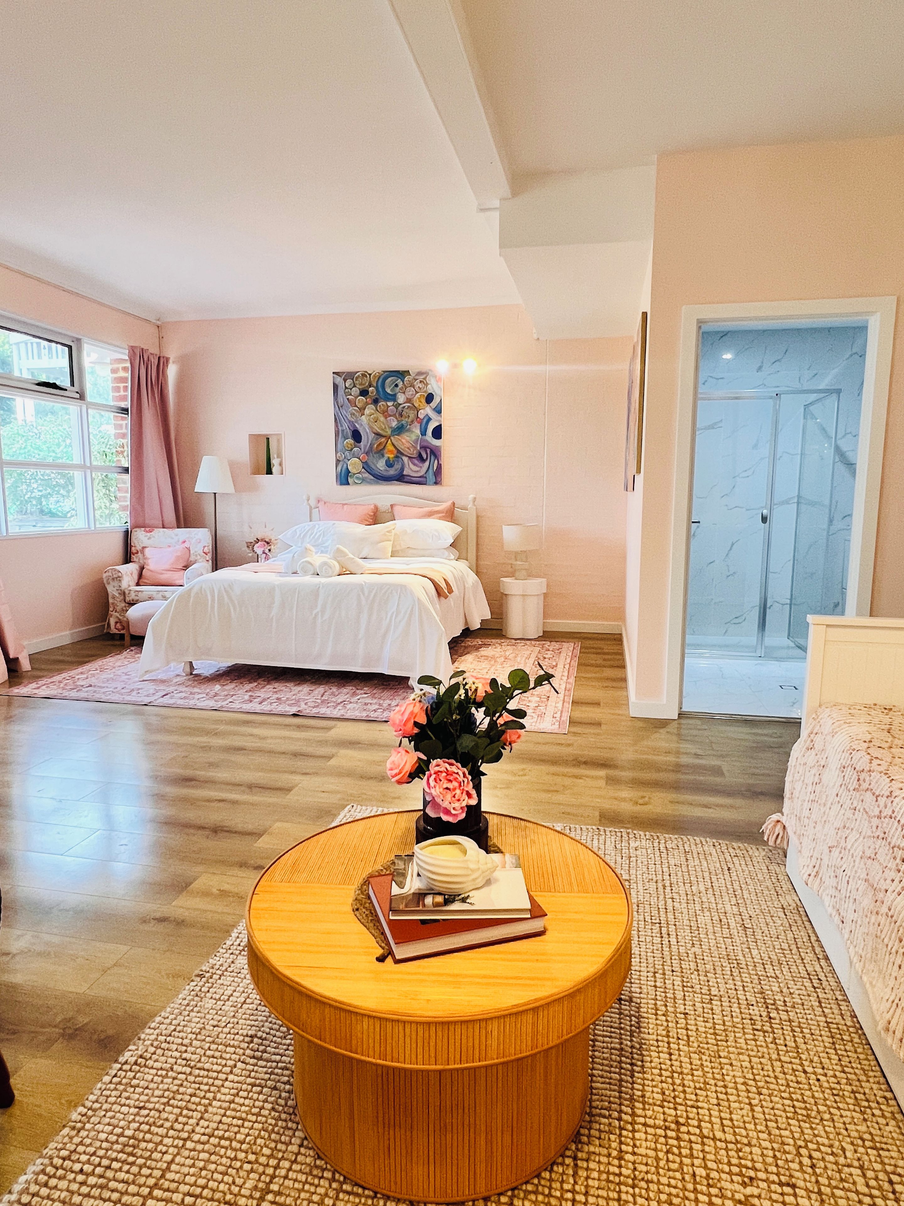 A bedroom with a bed, bedside lamps, a round wooden table with books and a vase of flowers, a window with curtains, and a door.