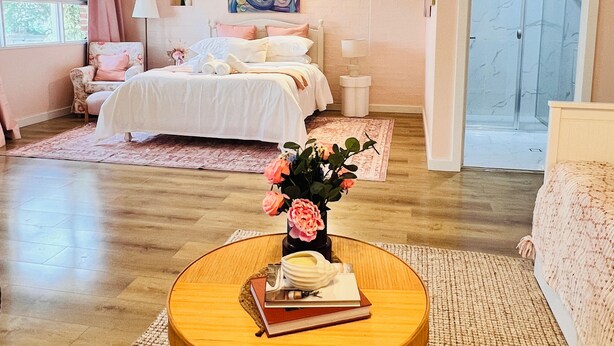 Quarto com cama, abajures, mesa redonda de madeira com livros e um vaso de flores, janela com cortinas e porta.