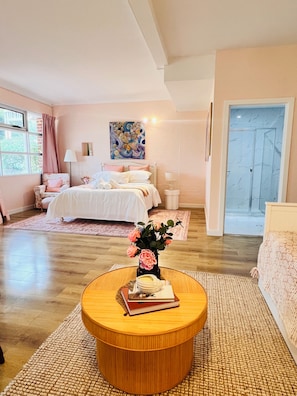 Un dormitorio con cama, lámparas de noche, una mesa redonda de madera con libros y un jarrón de flores, una ventana con cortinas y una puerta.