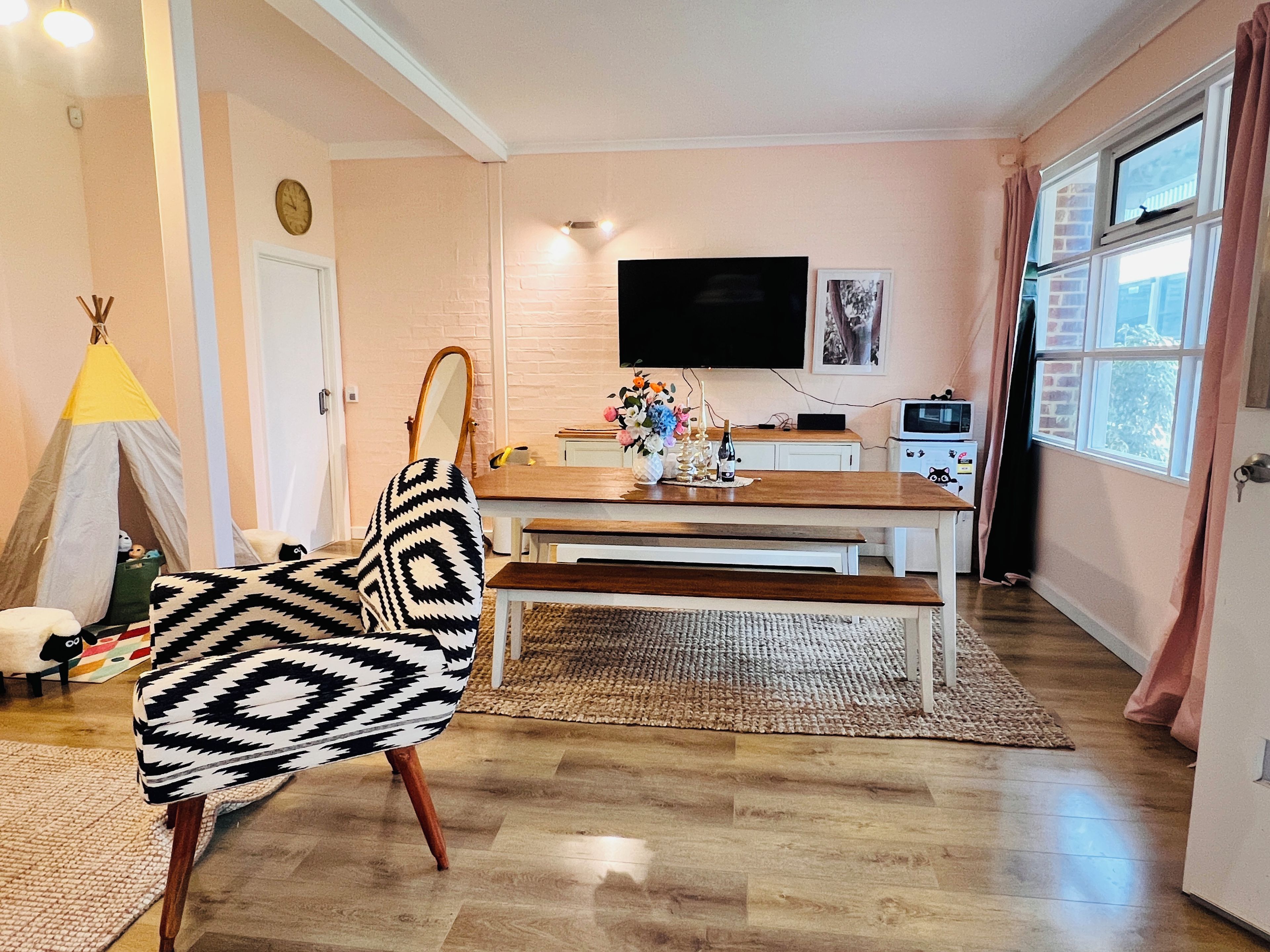 A living room with a wooden dining table, a sofa with a patterned cover, a television mounted on the wall, and a window with curtains.