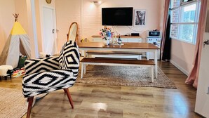 Uma sala de estar com uma mesa de jantar de madeira, um sofá com capa estampada, uma televisão fixada na parede e uma janela com cortinas.