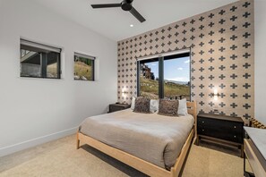 Ein Schlafzimmer mit Bett, Nachttisch, Kommode, Deckenventilator und einem Fenster mit Blick auf ein Haus und eine Landschaft.