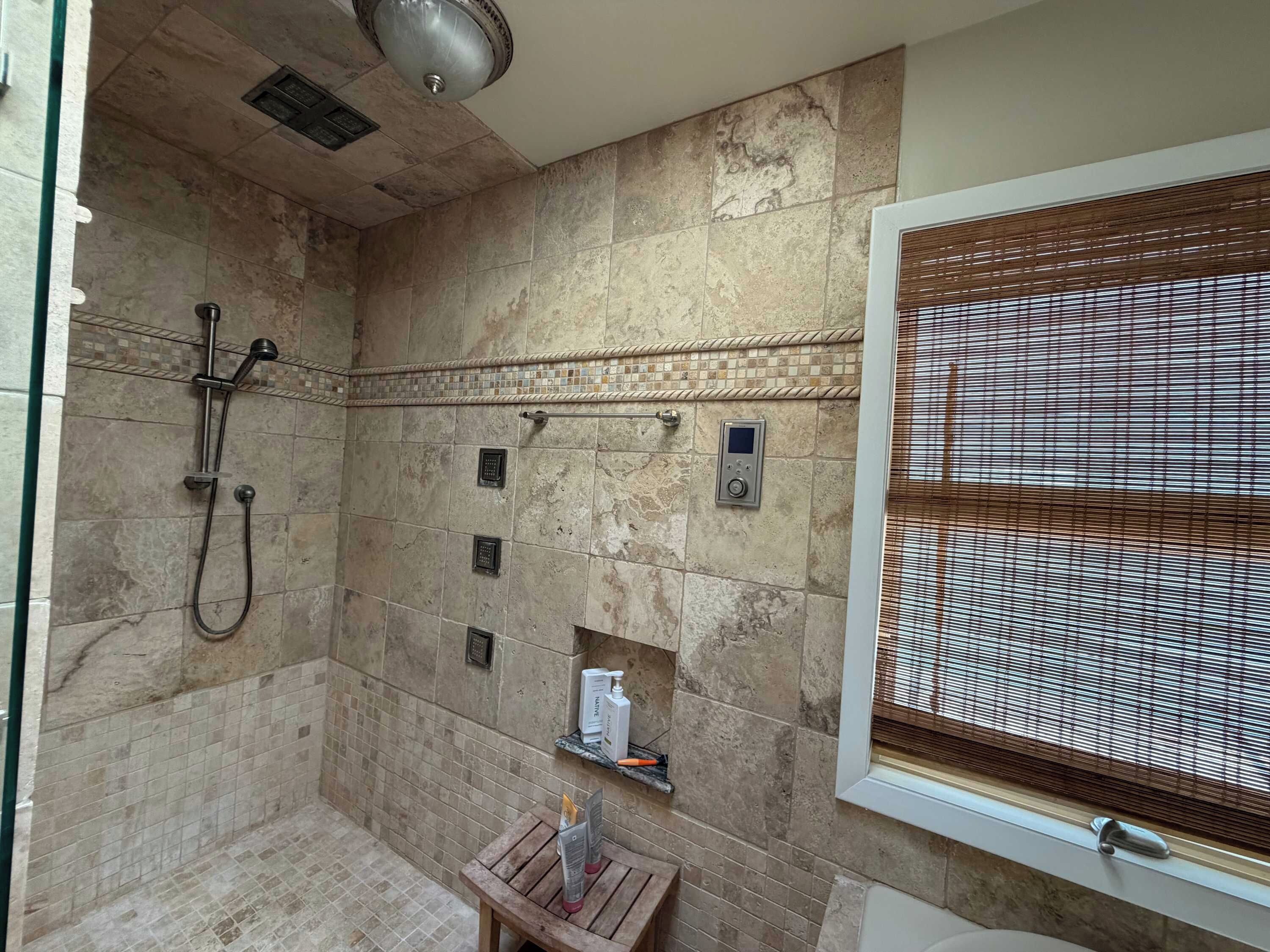A bathroom with a shower, a control panel, a window with blinds, and a small wooden table with toiletries.