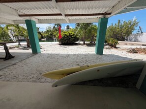 Ein überdachter Außenbereich mit einem Surfbrett, das an einer Wand lehnt, einer Bank und sandigem Boden.