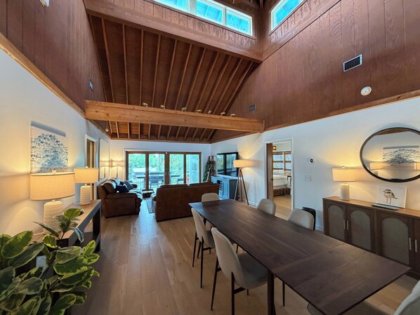 A spacious living area with a high ceiling, wooden beams, a dining table, and a view through large sliding doors to an outdoor deck. - Cypress Cove - Private Canal Front Retreat in Sugarloaf (Sugarloaf Key)