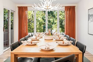 Ein Esszimmer mit einem großen Holztisch, der für eine Mahlzeit gedeckt ist, umgeben von Stühlen, mit Blick auf Grün durch große Fenster.