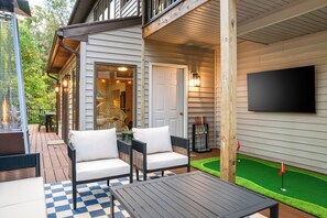 An outdoor patio area with a checkered floor, a table, two chairs, a TV mounted on the wall, and a small putting green. - Wild & Well - Red Light • Sauna • Gym • Hot Tub (McGaheysville)