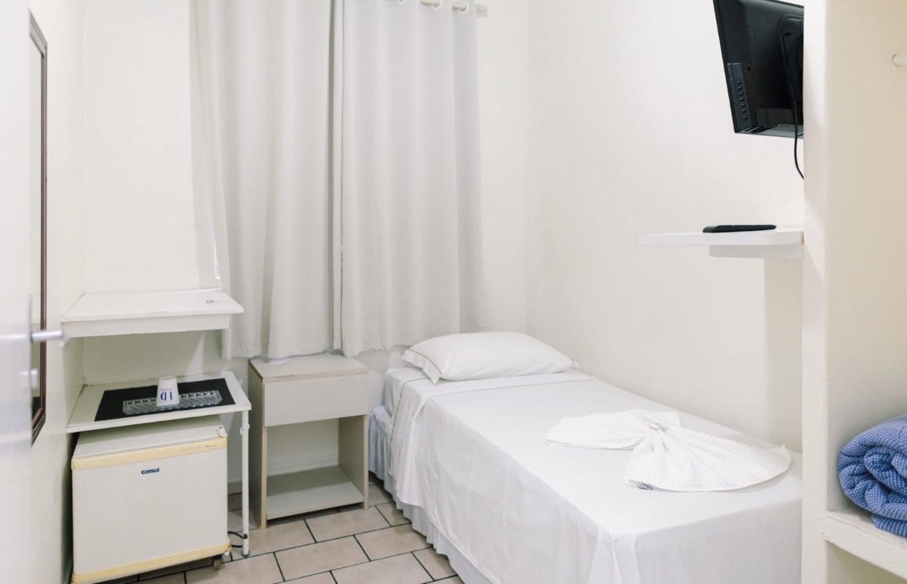A small, clean room with a single bed, a bedside table, a small cabinet, a TV mounted on the wall, and a curtain.