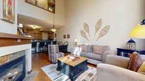 A spacious living room with a fireplace, a sofa, a coffee table, and a dining area with chairs. - Stone's Throw #16 | 3 Peaks Rentals (Sun Peaks)