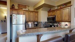 A kitchen with wooden cabinets, a stainless steel refrigerator, a microwave, and a granite countertop. - Stone's Throw #16 | 3 Peaks Rentals (Sun Peaks)