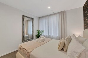 Une chambre d’hôtel avec un grand lit, un miroir, une plante et une fenêtre avec des rideaux.