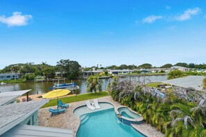 A pool area with lounge chairs and umbrellas, a small pond, and a view of a residential area with houses. - Canalfront 4BR with Pool, Dock & Kayaks (Cocoa Beach)