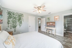 A bedroom with a large bed, a ceiling fan, a wall-mounted TV, and a bathroom visible through an open door. - Canalfront 4BR with Pool, Dock & Kayaks (Cocoa Beach)