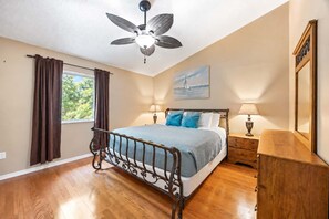 Ein Schlafzimmer mit einem Bett, einem Deckenventilator, einem Fenster mit Vorhängen, einem Gemälde und einem Nachttisch mit Lampe.