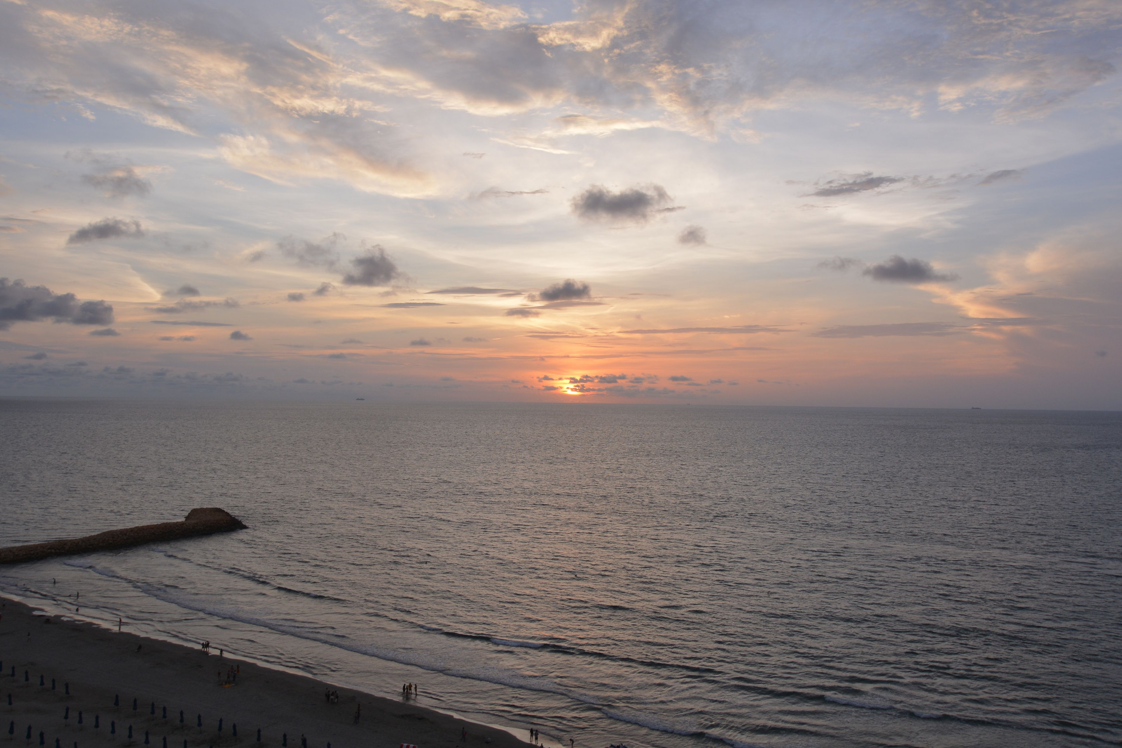 Una spiaggia con un molo che si estende verso il mare, onde che lambiscono dolcemente la riva e un tramonto sullo sfondo.