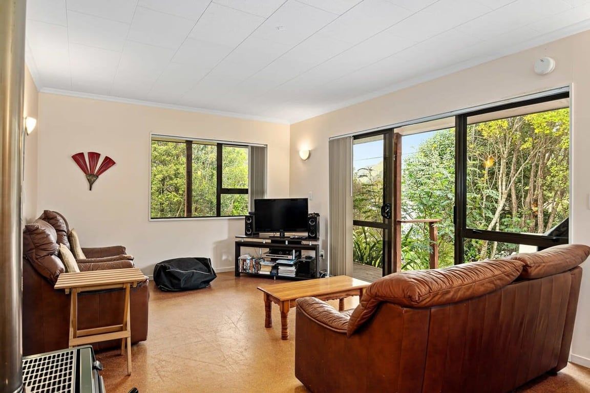 A living room with a brown leather sofa, a television on a stand, a wooden coffee table, and a large window with a view of trees.