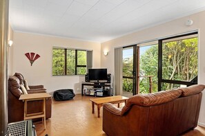 A living room with a brown leather sofa, a television on a stand, a wooden coffee table, and a large window with a view of trees.