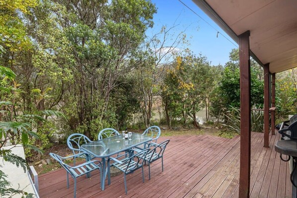 An outdoor patio with a glass table and chairs, surrounded by dense vegetation and a wooden deck.
