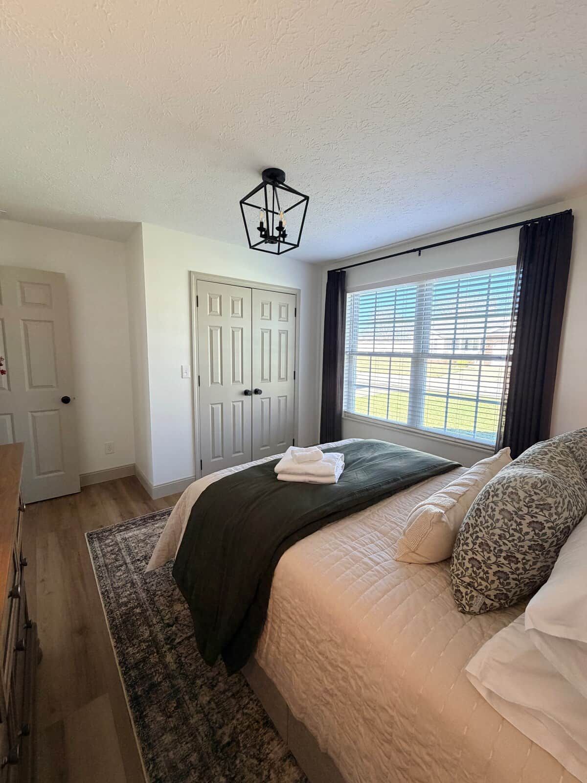 Quarto com cama, janela com persianas, porta e luminária suspensa.