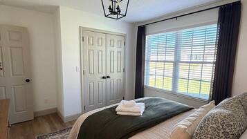 Quarto com cama, janela com persianas, porta e luminária suspensa.