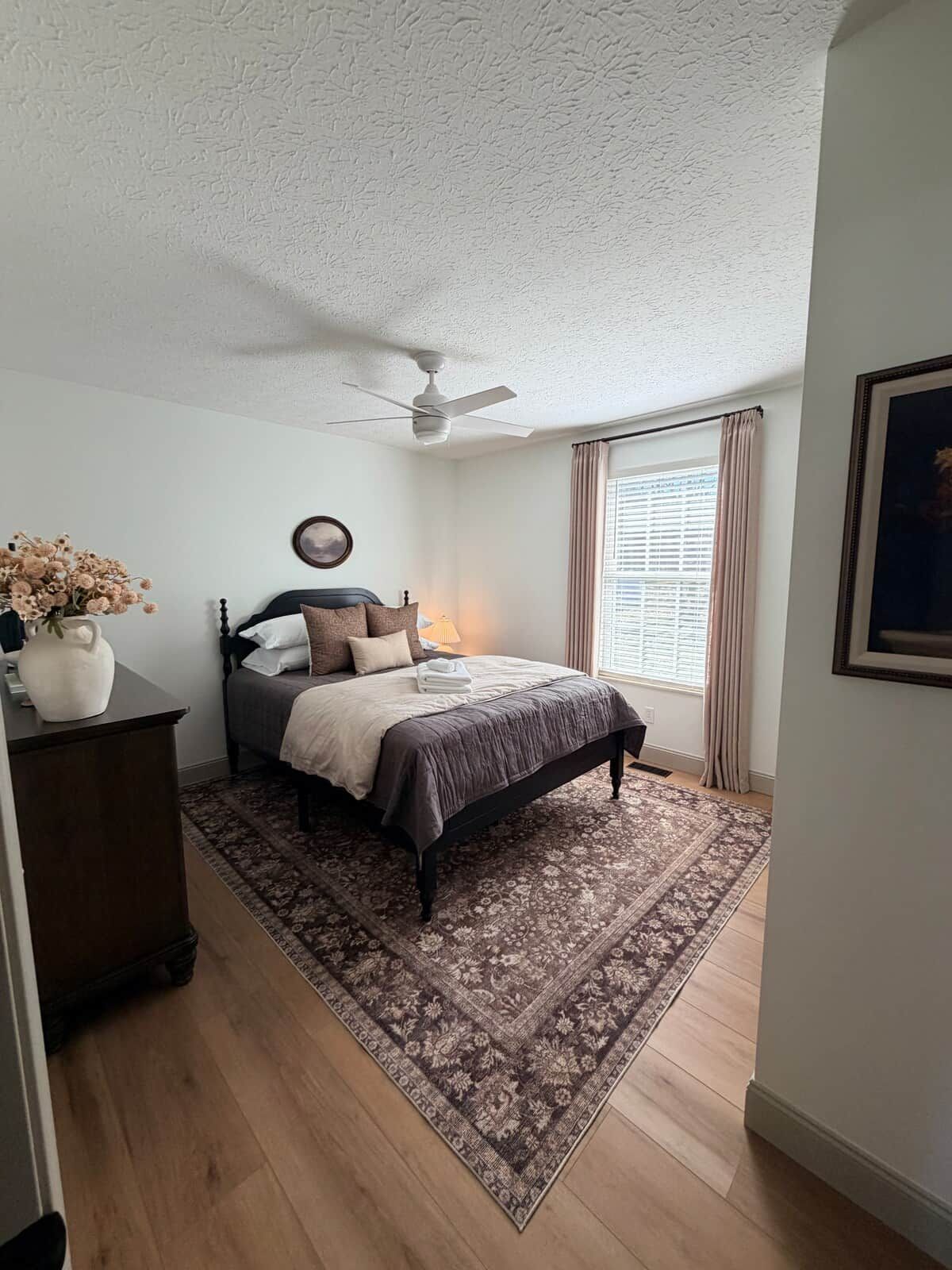 A bedroom with a bed, a ceiling fan, a window with curtains, and a nightstand with a vase.