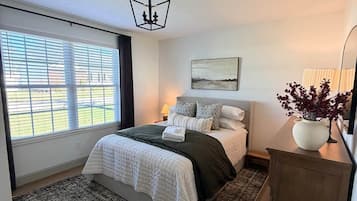 A bedroom with a bed, a large window, a nightstand, a lamp, a painting, and a cabinet.
