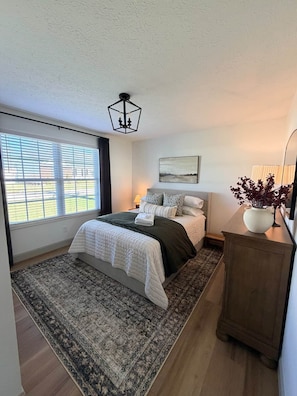 A bedroom with a bed, a large window, a nightstand, a lamp, a painting, and a cabinet.