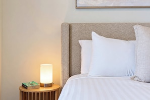 A neatly made bed with white bedding and pillows, a round wooden bedside table with a lamp, and a book titled 'Normal People' on the table. - Sea Vista at The One (Newcastle West)