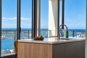 A modern kitchen with a view of the sea, a sink, and a bowl of fruit on the counter. - Sea Vista at The One (Newcastle West)