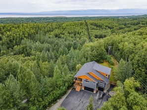 A two-story house with a garage, surrounded by dense forest, with a view of distant mountains. - The Chugach Retreat-Spacious Luxury, Views & Hot Tub, Sleeps 10, FlexibleBooking (Anchorage)