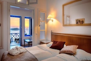 A hotel room with a bed, a nightstand, a lamp, and a view of the sea through a door. - Golden Grey Goose - Adults Only (Santorini)