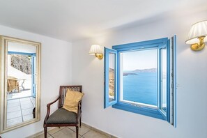 A room with a view of the sea, a chair with a cushion, and a mirror reflecting an outdoor area. - Golden Grey Goose - Adults Only (Santorini)