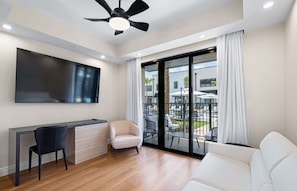 Una sala de estar moderna con televisor de pantalla plana, un escritorio, una silla, un sofá y un balcón con mobiliario exterior.