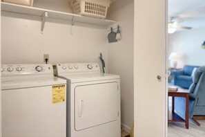 Uma área de lavanderia com máquina de lavar e secadora de abertura frontal, um cesto em uma prateleira e uma porta que dá para uma sala de estar.