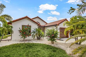 Une maison avec un toit en tuiles, une porte d’entrée et un petit jardin agrémenté de palmiers et de fleurs rouges.