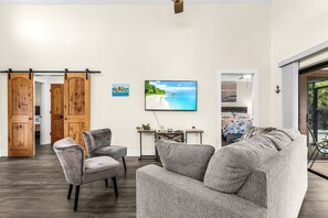 Ein Wohnzimmer mit einer großen grauen Couch, einem kleinen grauen Sessel, einer Holztür, die sich schieben lässt, einem an der Wand befestigten Fernseher und einem Tisch mit zwei Topfpflanzen.