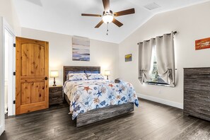 Une chambre avec une porte en bois, un lit recouvert d’une couvre-lit à motifs floraux, un ventilateur de plafond et une fenêtre avec des rideaux.