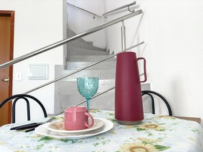 Ein Esstisch mit einer blumigen Tischdecke, einer rosa Tasse, einem roten Thermosbehälter und einem blauen Glas.