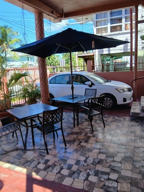 Un espacio al aire libre con mesas y sillas, un coche blanco estacionado cerca y un edificio al fondo.