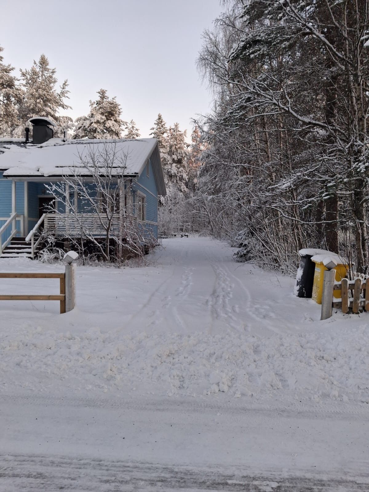 En snötäckt väg leder fram till ett hus med en synlig trappa och ett trästaket.