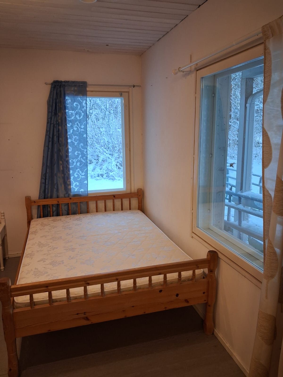Quarto pequeno com uma cama de solteiro, estrutura de madeira, janela com vista para uma paisagem nevada e cortina entreaberta.