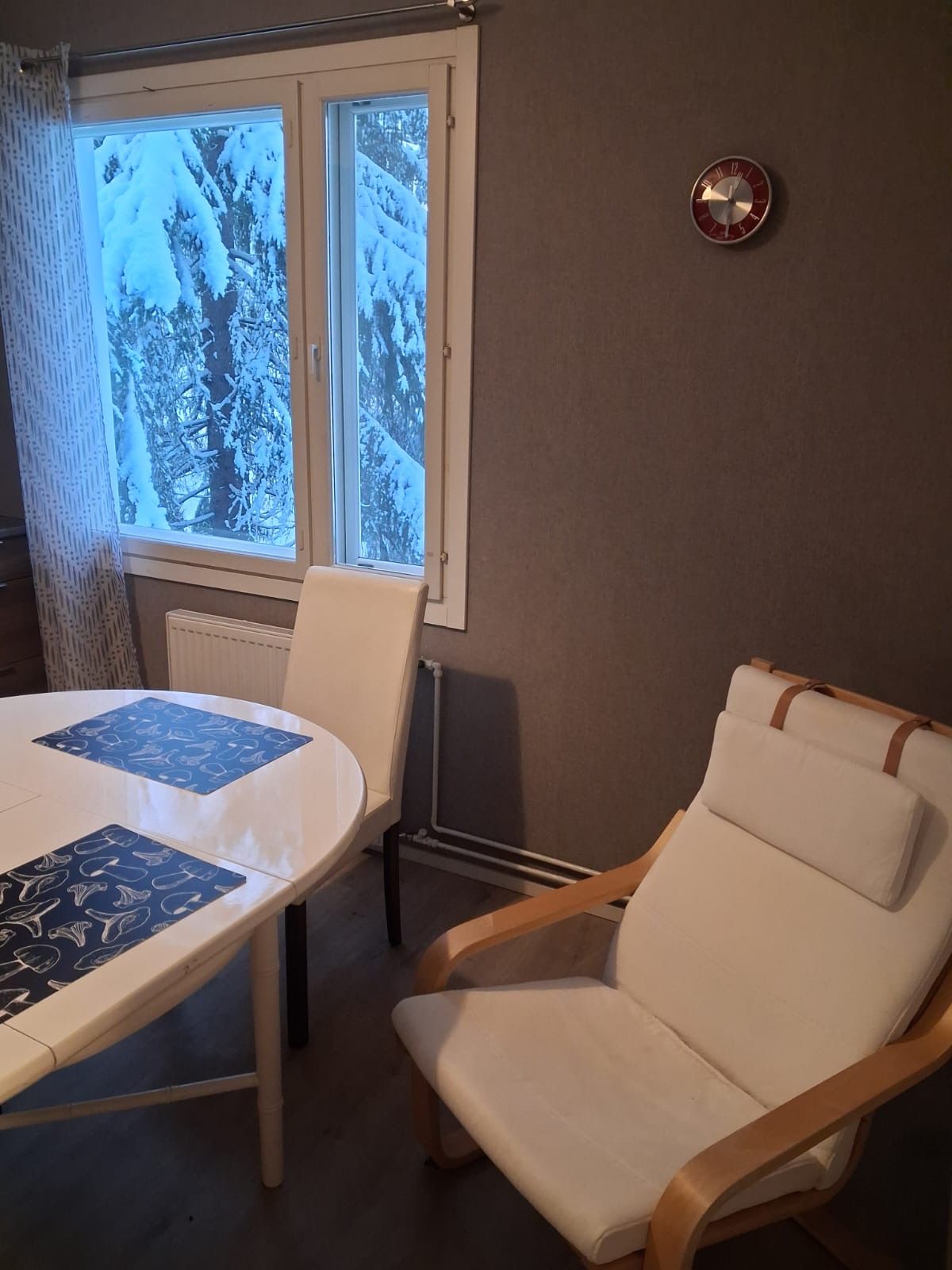 Ett rum med ett runt bord och en enda vit stol, ett fönster med utsikt över snötäckta träd och en väggklocka.