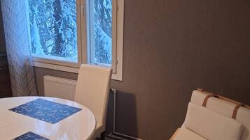 Ett rum med ett runt bord och en enda vit stol, ett fönster med utsikt över snötäckta träd och en väggklocka.