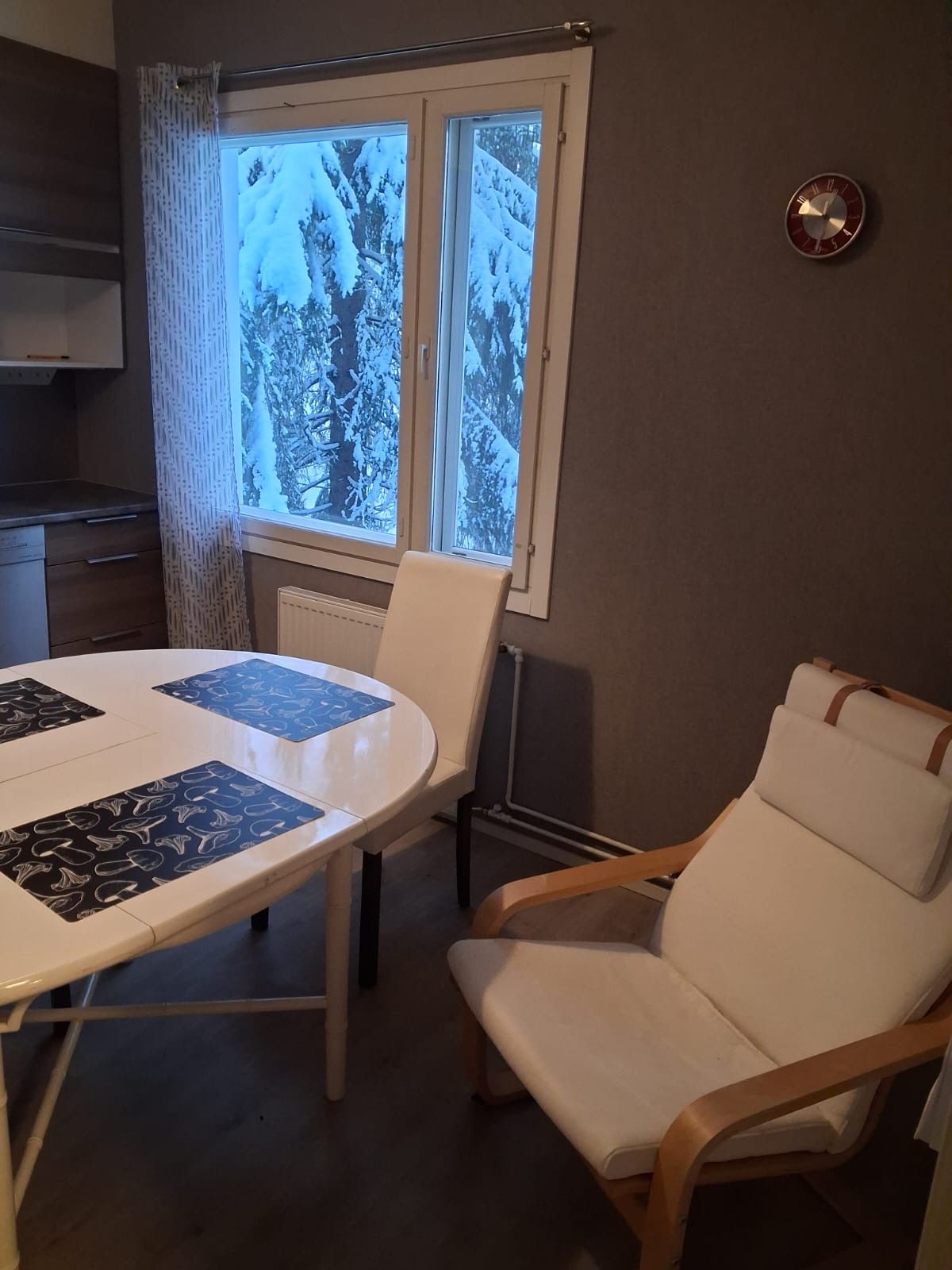 Huoneessa on pyöreä pöytä, valkoinen tuoli ja ikkuna, josta avautuu näkymä lumipeitteisiin puihin.