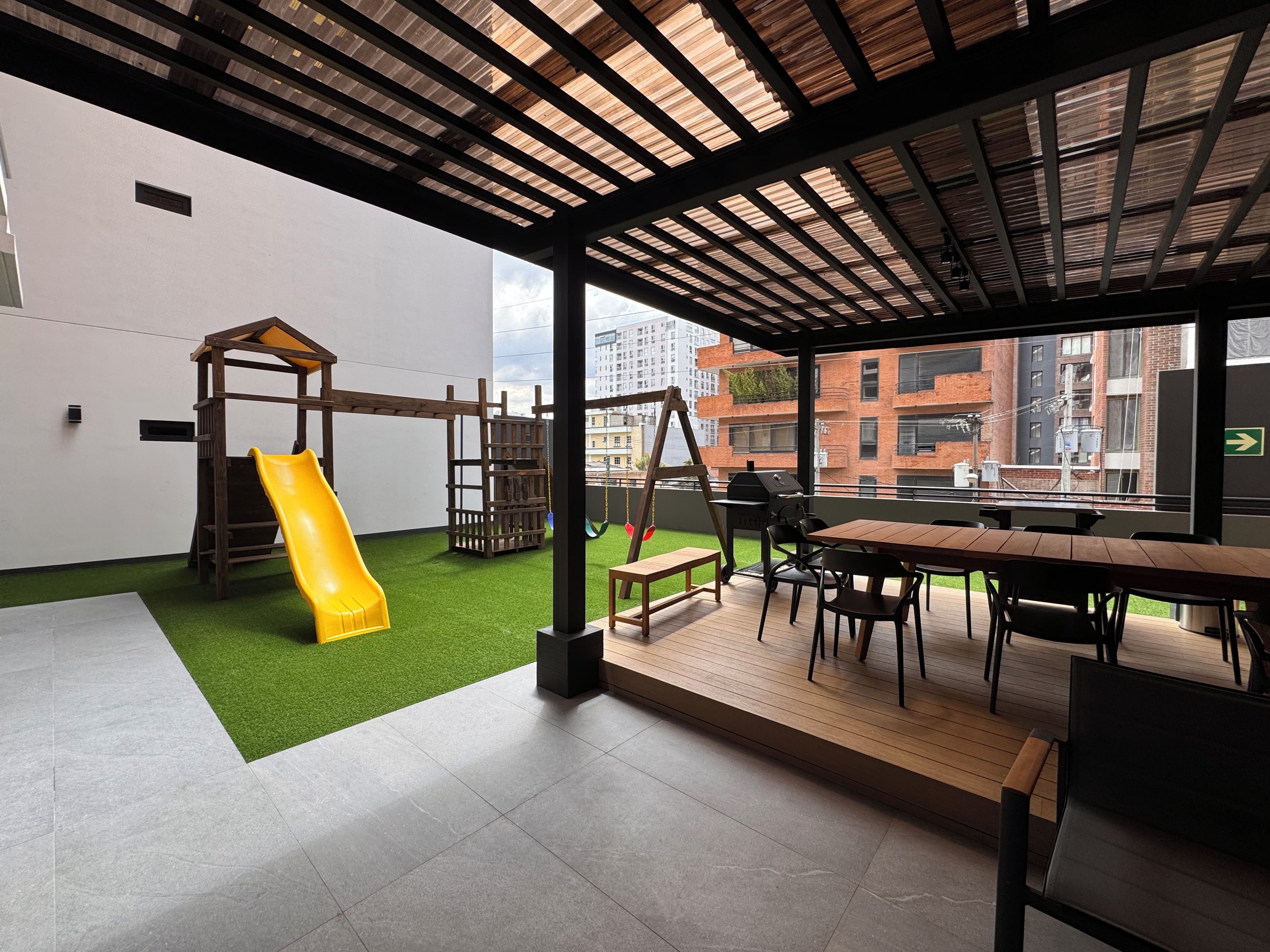 A children's play area with a slide and climbing structures, a patio with a table and chairs, and a view of buildings through large windows.