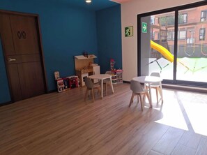 A room with a wooden floor, a blue wall, a door with a toilet symbol, a window with a view of a playground, and a table with chairs. - Aurea Luxury Apartment Z10 (Guatemala)